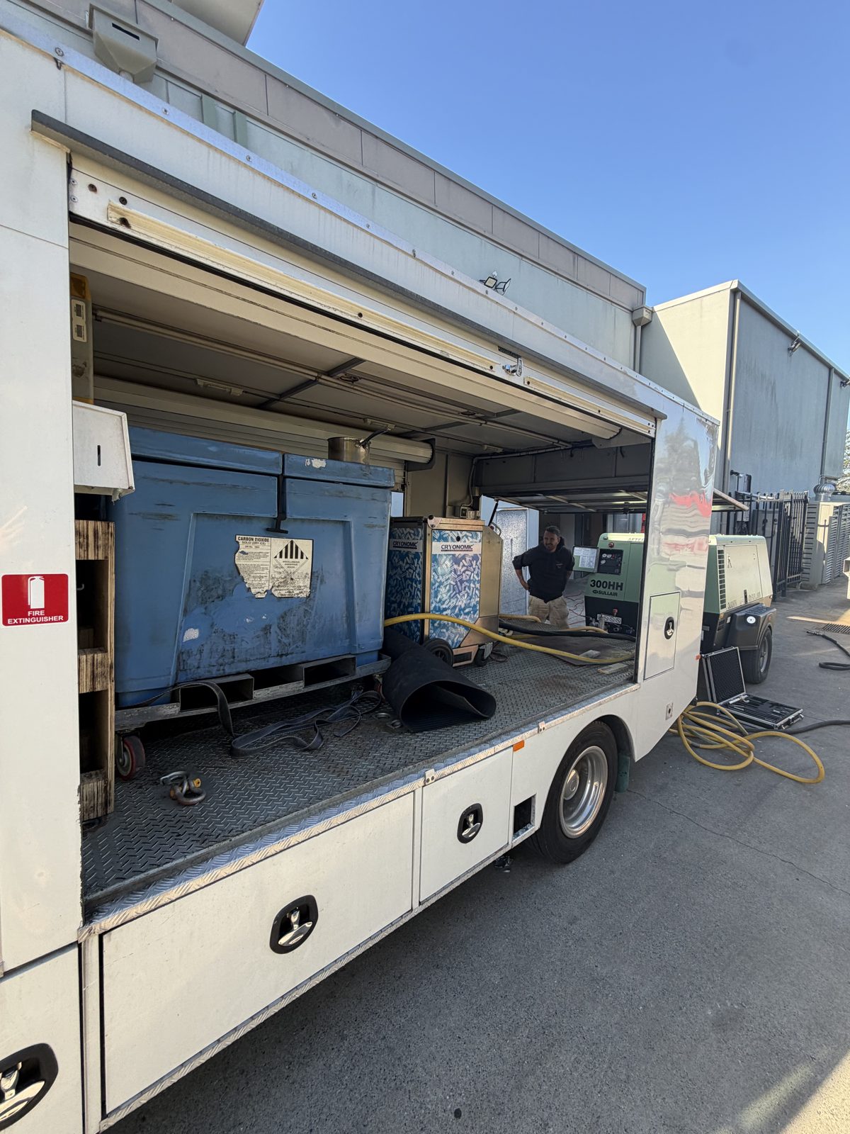 Dry Ice Blasters box truck on site at the golf club with roller door up — insulated pellet bin, compressor and Cryonomic unit inside, hoses run out