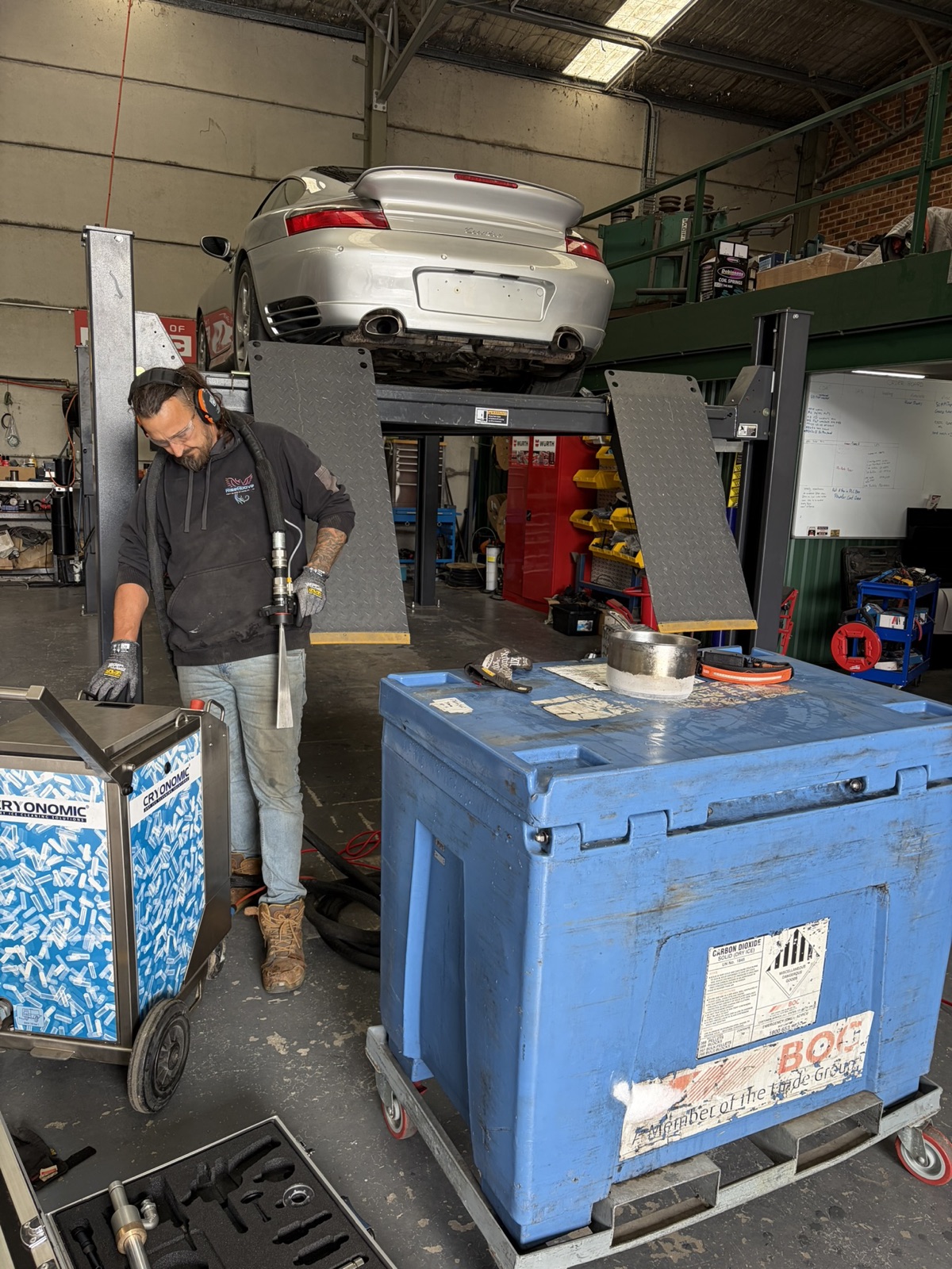 Operator preparing the Cryonomic dry ice blasting unit beside an insulated pellet bin, Porsche on the lift behind